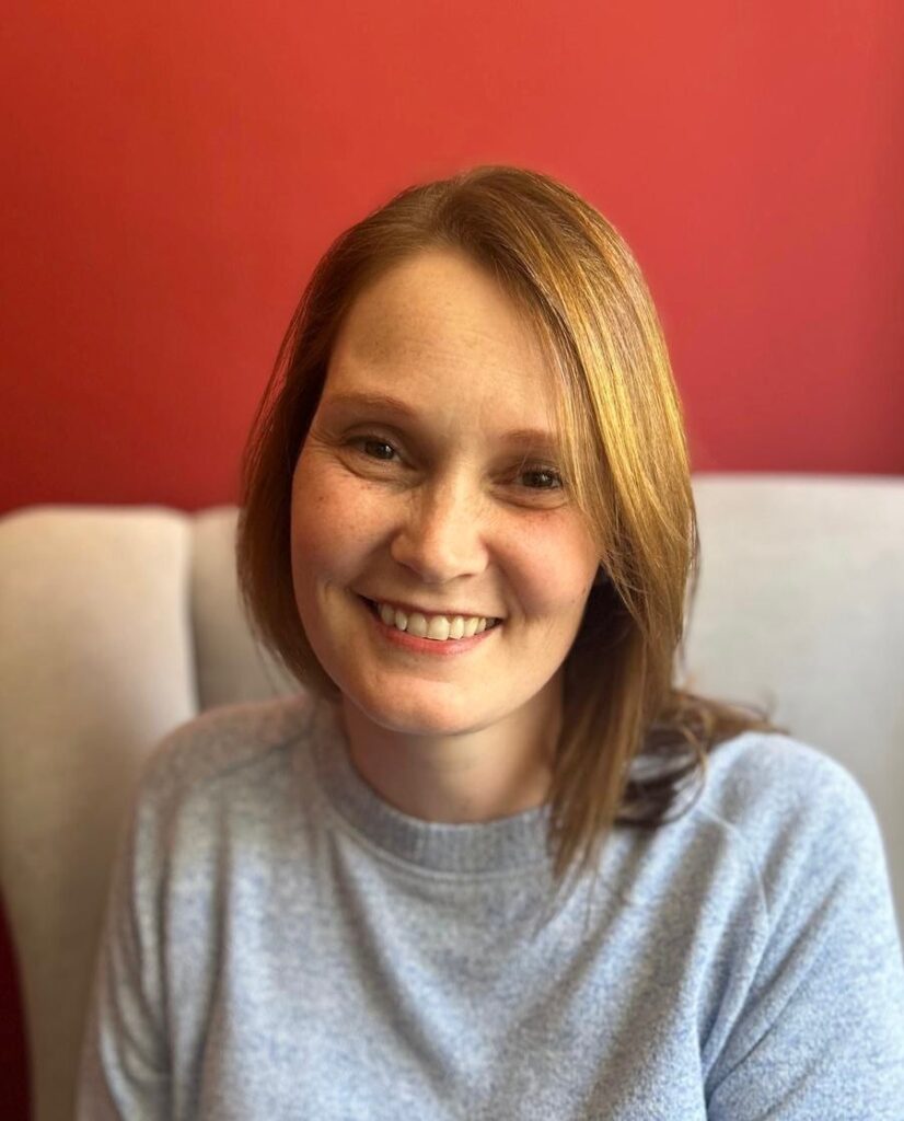 Founder - Rianne Garth - Lead Physiotherapist - Lady sat smiling on a white sofa with a red/burgundy wall behind.